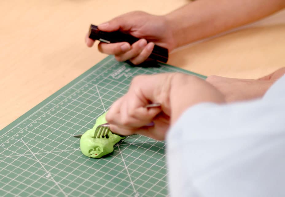 Person using a fork to shape green modeling clay on a cutting mat while holding a sculpting tool.